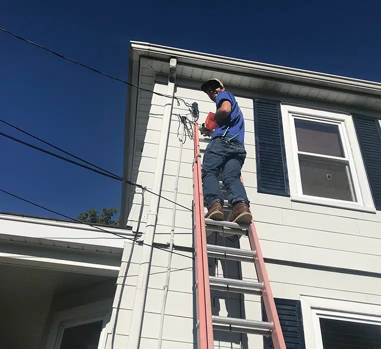 Electrician on ladder performing exterior service entrance work on Eastern Massachusetts home