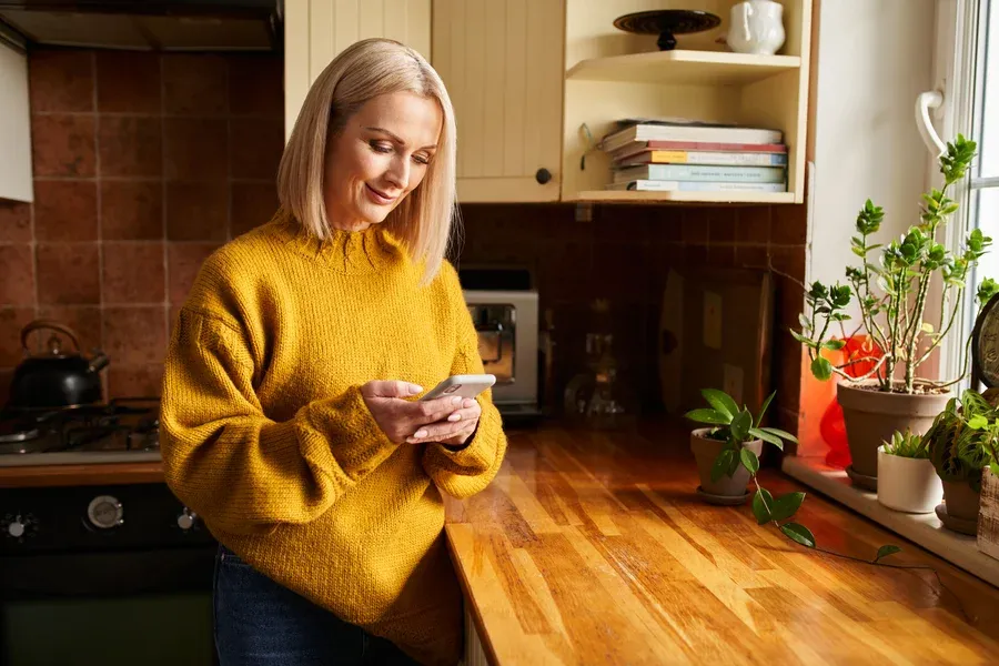 Woman in sweater smiling at her phone in a kitchen, signing up for Castle Electric SMS deals and tips in Eastern Massachusetts