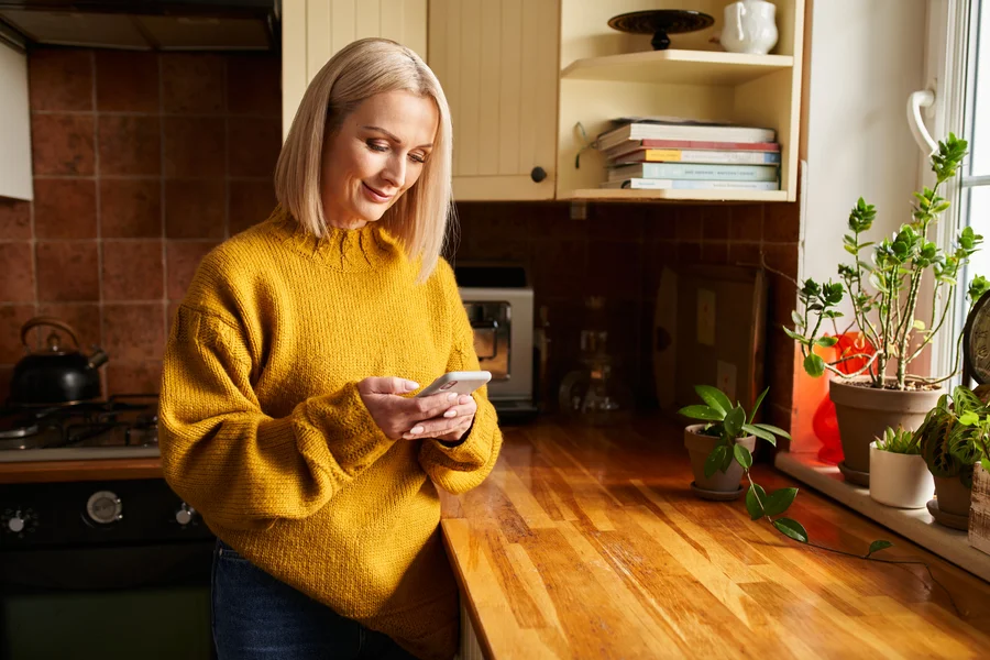 Woman in sweater smiling at her phone in a kitchen, signing up for Castle Electric SMS deals and tips in Eastern Massachusetts